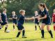 Un garçon de 8 ans apprend les bases du rugby avec une entraîneuse sur un terrain de gazon, entouré de jeunes coéquipiers dans une ambiance accueillante.
