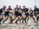 Une équipe masculine de rugby réalise une action de passe sur un terrain boueux et détrempé sous une forte pluie.
