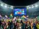 Supporters hommes et femmes agitant des drapeaux nationaux dans un stade de football européen illuminé la nuit, avec un grand écran lumineux aux formes abstraites.
