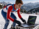 Jeune skieur alpin français en tenue de course consultant un ordinateur affichant des graphiques financiers abstraits, devant un paysage montagneux enneigé.